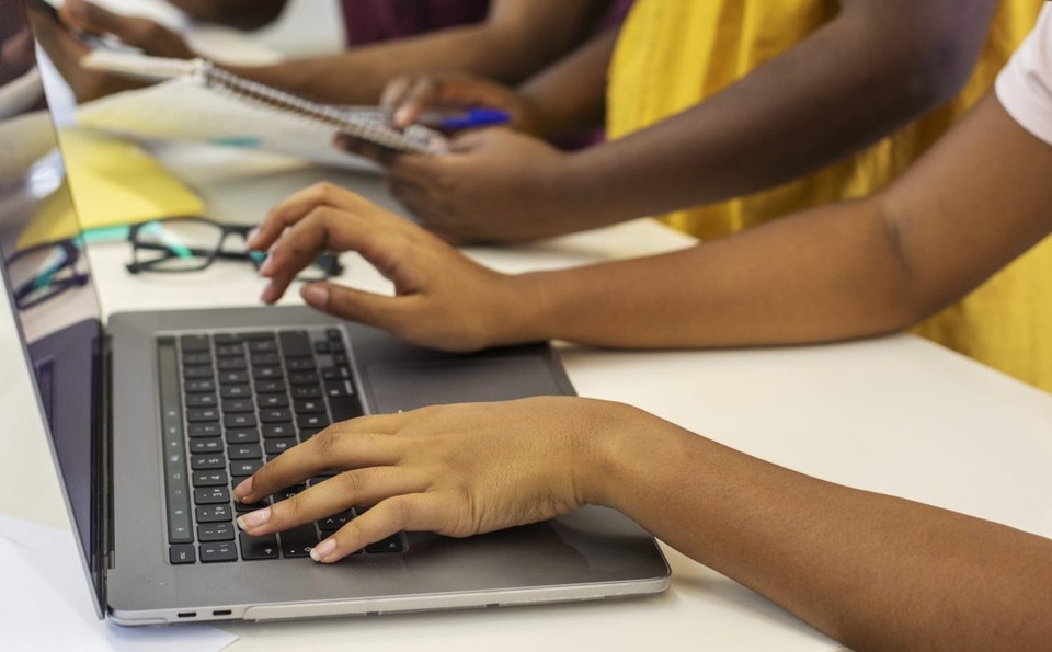 Mãos de jovens digitando em um notebook e escrevendo em um caderno sobre uma mesa